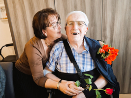 Eine Frau umarmt einen lächelnden älteren Mann mit einer Rose in der Hand.