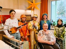 Eine Gruppe von sechs Personen feiert mit einer älteren Frau in einem Krankenhauszimmer.