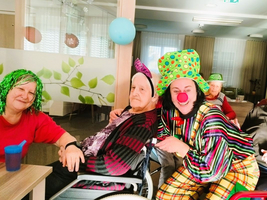 Ein Clown posiert fröhlich mit mehreren Seniorinnen in einem Gemeinschaftsraum.