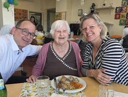 Drei Personen sitzen an einem Tisch mit einem Teller Essen. Eine ältere Frau in der Mitte lächelt, während zwei Erwachsene sich neben ihr platzieren und ebenfalls lächeln. Es gibt Gläser und einen Tisch mit einer gemusterten Tischdecke. Im Hintergrund sind andere Personen und Dekorationen zu sehen.