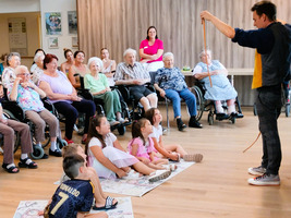 Ein Zauberer unterhält eine Gruppe von Senioren und Kindern in einem Gemeinschaftsraum.