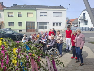Eine Gruppe älterer Menschen, einige im Rollstuhl, stehen vor einem Gebäude. Sie lächeln und posieren gemeinsam, während bunte Bänder und Zweige im Vordergrund sichtbar sind. Im Hintergrund sind Wohnhäuser und ein Auto zu sehen.