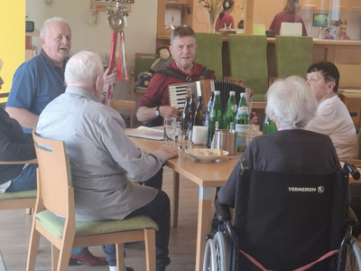 Senioren sitzen in einem Raum, während jemand Akkordeon spielt und sie unterhalten.