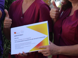 Zwei Personen halten ein Pflegezertifikat und zeigen Daumen hoch.
