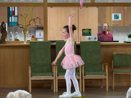 Ein Mädchen in einem rosa Ballettkleid führt einen Tanz auf einer Kiste vor einem Publikum auf. Im Hintergrund sind grüne Stühle und ein Empfangstresen zu sehen. Über der Bühne hängen bunte Buchstaben. Zwei ältere Frauen im Publikum beobachten die Darbietung.