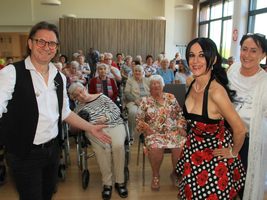 Eine fröhliche Veranstaltung in einem Seniorenheim mit einem Sänger und zwei Tänzerinnen im Vordergrund. Im Hintergrund sitzen viele ältere Menschen, einige im Rollstuhl, und genießen die Darbietung. Die Atmosphäre ist lebhaft und einladend.