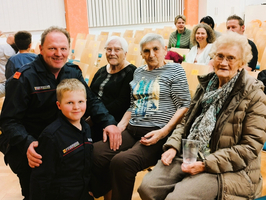 Eine Gruppe von fünf Personen sitzt beim Event in einem Saal. Zwei ältere Frauen, ein älterer Mann in Uniform, ein Junge und eine weitere ältere Frau posieren freundlich. Im Hintergrund sind andere Menschen zu sehen, die ebenfalls an dem Event teilnehmen.