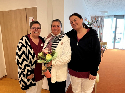 Drei Frauen stehen lächelnd zusammen, eine hält einen Blumenstrauß in der Hand.