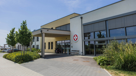 Eingang zu einem modernen Gebäude mit großen Fenstern und einem großen roten Kreuzsymbol an der Fassade. Vor dem Eingangsbereich befinden sich Bäume und eine blühende Bepflanzung. Der Weg ist gepflastert und führt zum Eingang.