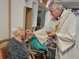Ein Priester reicht einer älteren Dame eine religiöse Gabe in einem Gemeinschaftsraum.