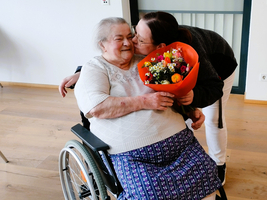 Eine ältere Frau in einem Rollstuhl erhält einen Blumenstrauß und umarmt eine Person.