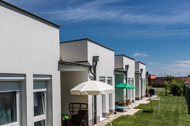 Moderne Wohnhäuser mit Gärten und Sonnenschirmen in ländlicher Umgebung.