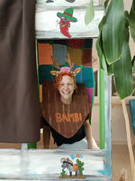Frau mit rotem Haar und Antlers steht lächelnd in einem bunten Rahmen. Sie trägt ein Schild mit der Aufschrift 
