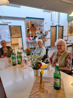 Drei ältere Frauen sitzen an einem Tisch mit Blumen und Getränken.