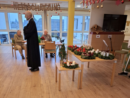 Ein Mann in Robe spricht zu älteren Menschen in einem Gemeinschaftsraum.
