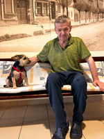 Ein älterer Mann sitzt auf einer Bank und hat einen kleinen Hund mit einem Geschirr neben sich. Beide schauen in die Kamera, während im Hintergrund eine historische Straßenszene zu sehen ist. Der Mann trägt ein grünes Shirt und eine schwarze Hose.