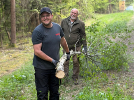 Zwei Männer stehen in einem Waldweg. Der vordere Mann trägt ein Holzstück und trägt Handschuhe. Der hintere Mann lächelt und ist in einem hellen Pullover gekleidet. Um sie herum sind grüne Pflanzen und Bäume.