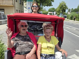 Drei Personen in einer Fahrrad-Rikscha im Freien, zwei ältere Frauen und eine Fahrerin.