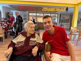 Junger Mann sitzt neben älterer Dame in einem Pflegeheim, beide lächeln.