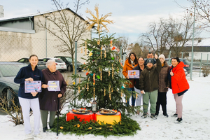 Gruppe von Menschen steht um einen festlich geschmückten Weihnachtsbaum im Schnee.