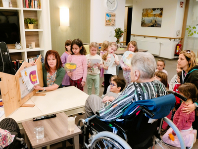Eine Gruppe von Kindern steht vor einer älteren Person im Rollstuhl, die an einer Präsentation teilnimmt. Eine Frau zeigt eine Zeichnung an einem Holzständer. Die Kinder sind aufmerksam und interessiert. Das Setting ist hell und freundlich, mit Möbeln im Hintergrund.