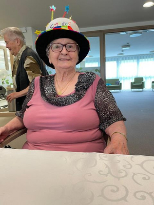 Eine ältere Frau mit Geburtstagskappe sitzt lächelnd an einem Tisch.