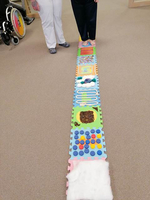 Zwei Personen stehen neben einem sensorischen Teppich mit verschiedenen Materialien.