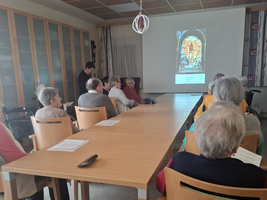 Eine Gruppe älterer Menschen sitzt an einem langen Tisch in einem Raum. Ein Mann steht vorne und präsentiert auf einer Leinwand mit einem bunten Bild. Die Teilnehmenden scheinen aufmerksam zuzuhören.