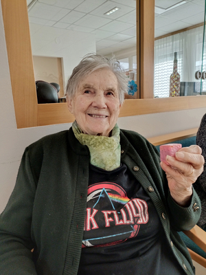 Eine lächelnde ältere Frau trägt ein T-Shirt von Pink Floyd und hält ein kleines Objekt.