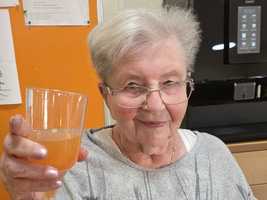 Eine ältere Frau hebt ein Glas mit einem orangefarbenen Getränk zum Anstoßen.