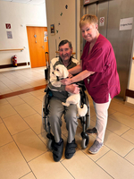Ein älterer Mann sitzt im Rollstuhl und hält einen kleinen Hund auf seinem Schoß. Neben ihm steht eine Frau in einem roten Oberteil, die freundlich lächelt. Im Hintergrund ist ein Aufzug und eine offene Tür sichtbar.