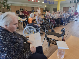 Eine Gruppe älterer Menschen sitzt in einem Gemeinschaftsraum, einige in Rollstühlen.