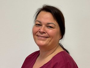 Lächelnde Frau mit braunen Haaren in einem burgunderfarbenen Oberteil vor hellem Hintergrund.