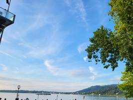 Menschen stehen an einem Gewässer unter einem blauen Himmel mit Wolken.