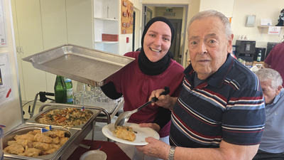 Eine lächelnde Mitarbeiterin in einem roten Oberteil und Kopftuch serviert einem älteren Mann mit einem gestreiften Hemd Essen aus einem Buffett. Der Mann hält einen Teller mit Speisen, während er begeistert in die Kamera schaut. Im Hintergrund sind weitere Essensangebote sichtbar.