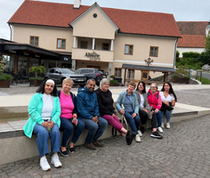 Eine Gruppe von neun Personen sitzt auf einer Steinbank vor einem Gebäude mit rotem Dach und dem Schriftzug 