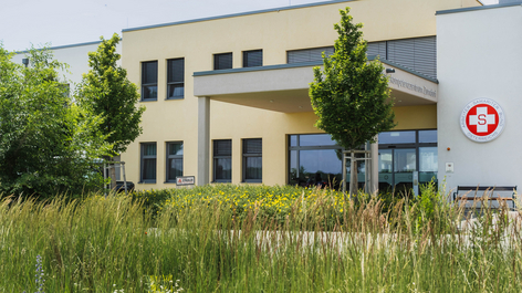 Außenansicht eines modernen Gebäudes mit großen Fenstern und einem Eingangsbereich, umgeben von grünen Bäumen und Wiesen. An der Fassade ist ein rotes Logo sichtbar. Der Gesamtstil ist freundlich und einladend.