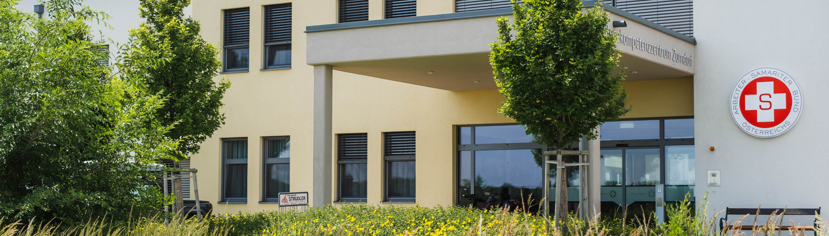 Außenansicht eines modernen Gebäudes mit großen Fenstern und einem Eingangsbereich, umgeben von grünen Bäumen und Wiesen. An der Fassade ist ein rotes Logo sichtbar. Der Gesamtstil ist freundlich und einladend.