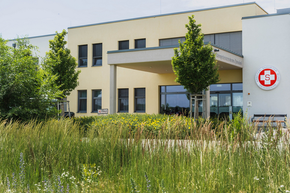 Außenansicht eines modernen Gebäudes mit großen Fenstern und einem Eingangsbereich, umgeben von grünen Bäumen und Wiesen. An der Fassade ist ein rotes Logo sichtbar. Der Gesamtstil ist freundlich und einladend.