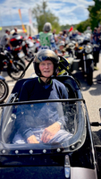 Ein älterer Mann sitzt in einem Motorradgespann und trägt einen Helm. Im Hintergrund sind zahlreiche Motorräder und Menschen auf einer Veranstaltung zu sehen. Der Himmel ist blau mit einigen Wolken.