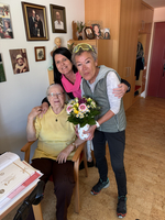 Drei Frauen stehen in einem Wohnraum. Eine ältere Frau sitzt auf einem Stuhl und hält einen Blumenstrauß in einer Vase. Zwei jüngere Frauen stehen neben ihr und strahlen. Hintergrund mit Bildern an der Wand.