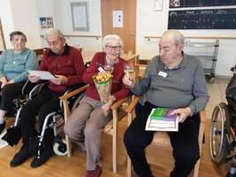 Vier ältere Menschen sitzen in einem Raum, einer hält Blumen und ein Glas.