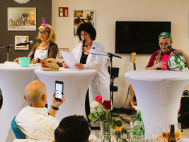 Drei Frauen in Kostümen halten eine unterhaltsame Show an Stehtischen.