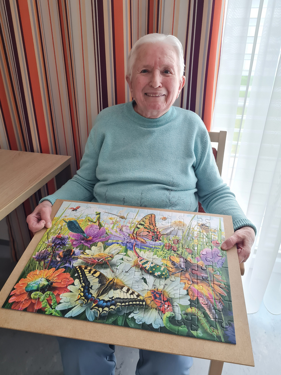 Ein älterer Mann mit weißem Haar sitzt an einem Tisch und lächelt, während er ein fertiges Puzzle mit bunten Schmetterlingen und Blumen in den Händen hält. Im Hintergrund sind gestreifte Vorhänge und ein Fenster zu sehen.
