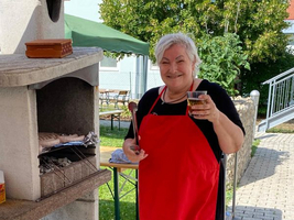 Eine Frau mit rotem Schürze hält ein Getränk und steht neben einem Grill.
