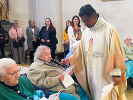 Eine Gruppe von Menschen in einem Kirchenraum. Ein Geistlicher überreicht einer älteren Frau ein Papier. Im Hintergrund sitzen weitere ältere Menschen, einige von ihnen haben Decken umgelegt. Die Atmosphäre wirkt feierlich und respektvoll.