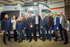 Grupp portrait vor einem Krankenwagen, bestehend aus mehreren Erwachsenen in Arbeitskleidung.
