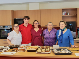 Fünf Personen stehen hinter einem Tisch, der mit verschiedenen Backwaren und Desserts gedeckt ist. Die Szenen spielen in einer modernen Küche, und die Gruppe lächelt in die Kamera. Im Hintergrund sind Küchenschränke und Geräte sichtbar.