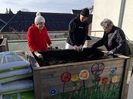 Drei Personen arbeiten zusammen an einem Hochbeet auf einem Balkon. Sie fügen Erde hinzu und holen gemeinsam Pflanzen hervor. Im Hintergrund sind Säcke mit Blumenerde gestapelt und die umliegenden Gebäude sind teilweise sichtbar.