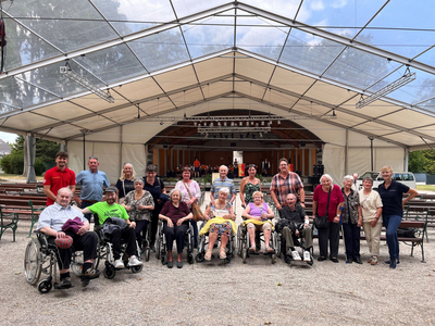 Eine Gruppe von Senioren, darunter mehrere Rollstuhlfahrer, steht unter einem großen Zelt. Im Hintergrund ist eine kleine Bühne sichtbar. Die Personen lächeln und posieren freundschaftlich für das Foto.